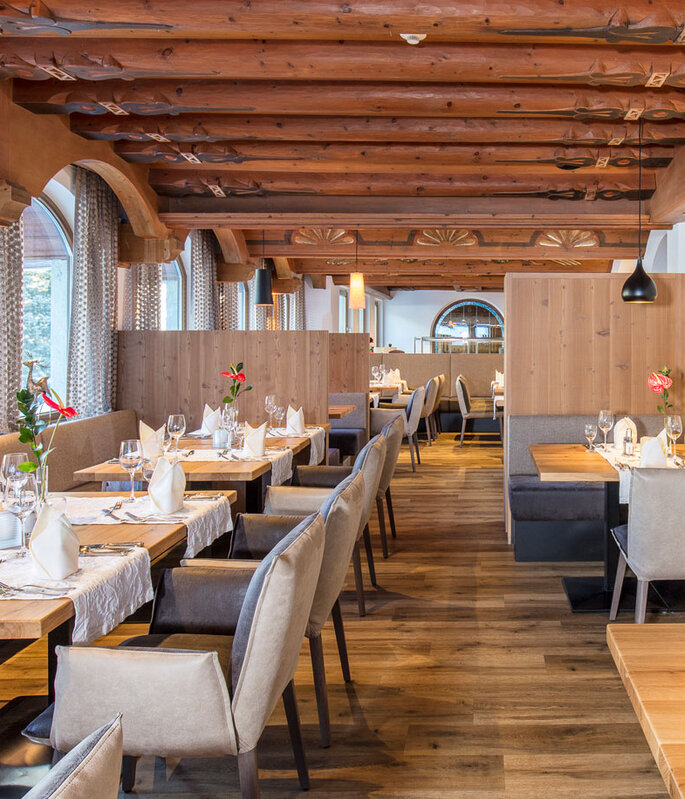 Ein elegantes Restaurant mit Holzbalken, großen Bogenfenstern, gepflegt gedeckten Tischen und gepolsterten Stühlen. Der Raum ist von natürlichem Licht durchflutet, und auf jedem Tisch stehen Blumen, was eine warme und einladende Atmosphäre schafft.