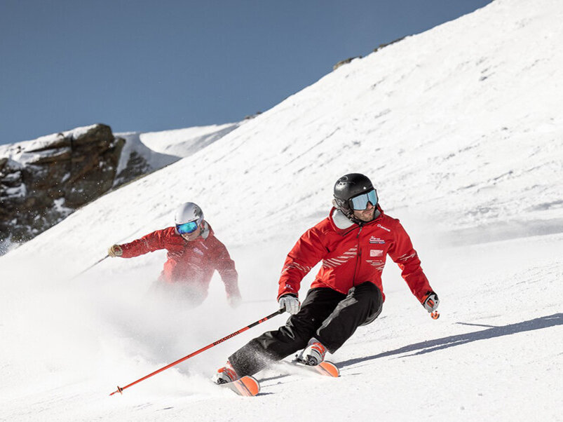 Zwei Skifahrer in roten Jacken und Helmen fahren unter strahlend blauem Himmel einen verschneiten Berghang hinunter, wobei hinter ihnen Schneewolken aufwirbeln.
