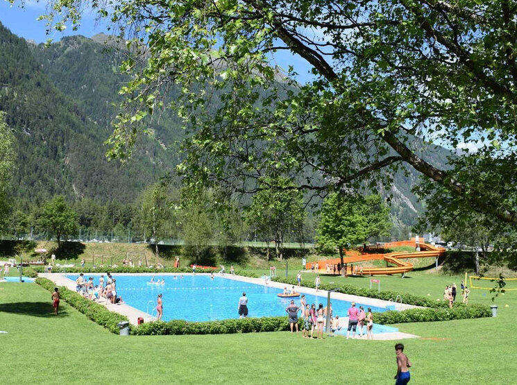 Die Menschen genießen einen sonnigen Tag an einem Freibad, das von grünem Rasen, Bäumen und Bergen umgeben ist. Einige schwimmen, entspannen sich auf ihren Handtüchern oder spielen Volleyball.