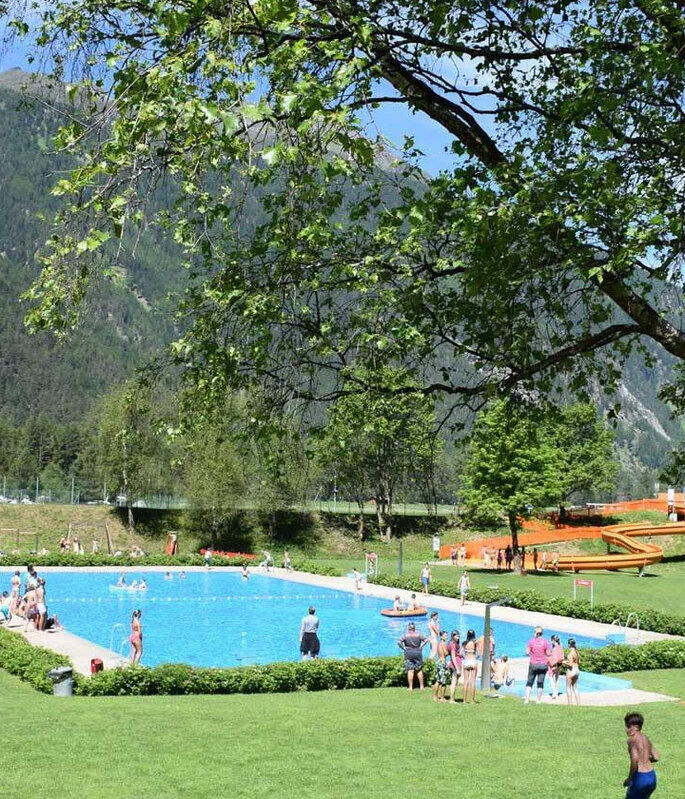 Die Menschen genießen einen sonnigen Tag an einem Freibad, das von grünem Rasen, Bäumen und Bergen umgeben ist. Einige schwimmen, entspannen sich auf ihren Handtüchern oder spielen Volleyball.