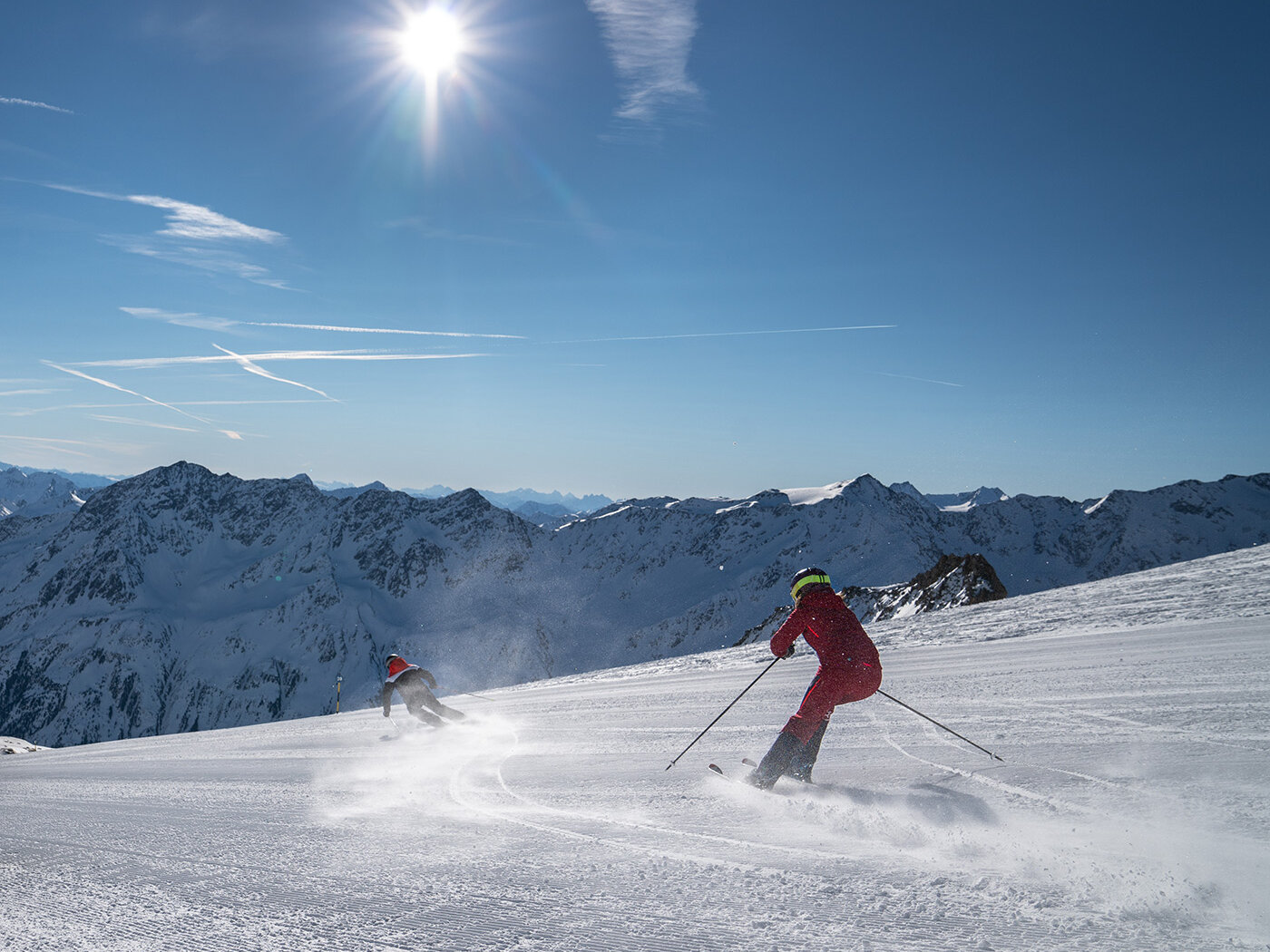 Zwei Skifahrer in bunten Outfits gleiten unter strahlendem Sonnenschein einen verschneiten Berghang hinunter, im Hintergrund sind schneebedeckte Gipfel und ein strahlend blauer Himmel zu sehen.