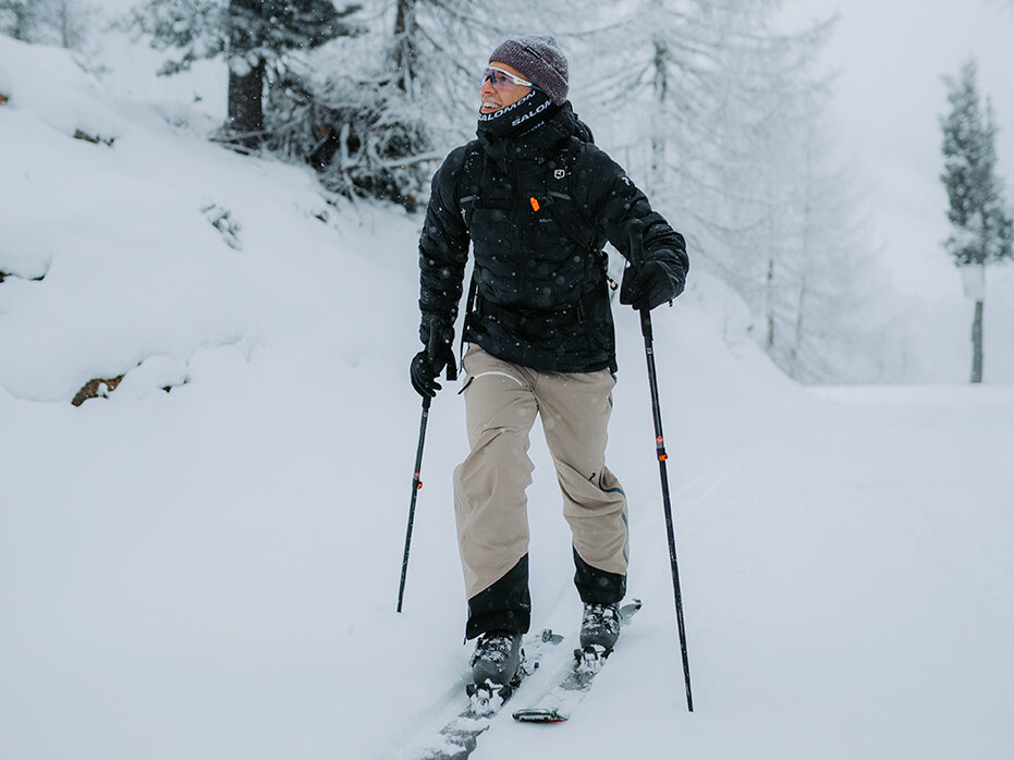 Eine Person in Winterkleidung geht mit Tourenski bergauf durch einen verschneiten Wald und stützt sich dabei auf ihre Skistöcke. Schnee bedeckt den Boden und die Bäume und schafft so eine friedliche Winterlandschaft.