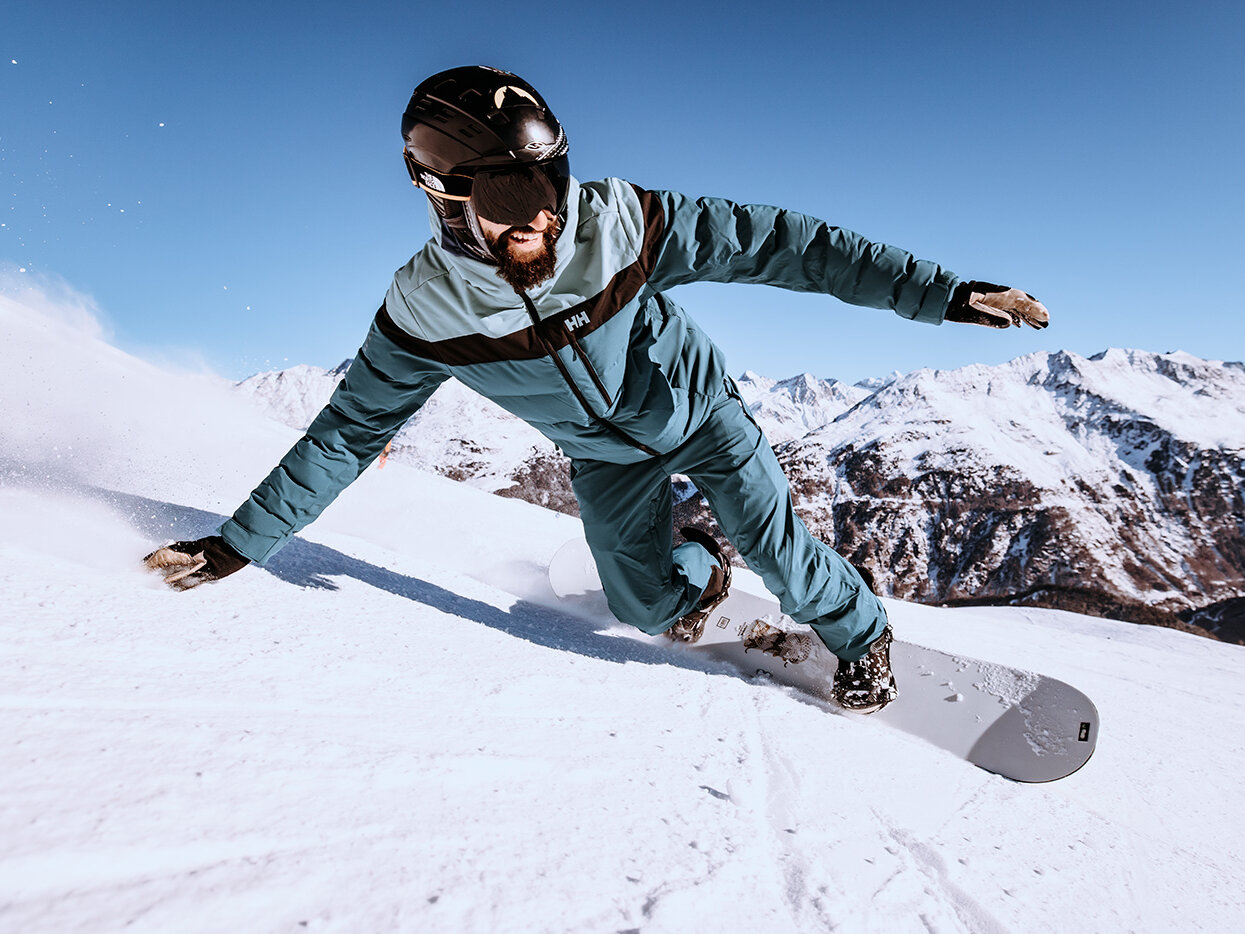 Eine Person fährt in einem blau-grauen Schneeanzug, mit Helm und Skibrille auf einem verschneiten Berghang Snowboard. Im Hintergrund sind schneebedeckte Berge und ein strahlend blauer Himmel zu sehen.