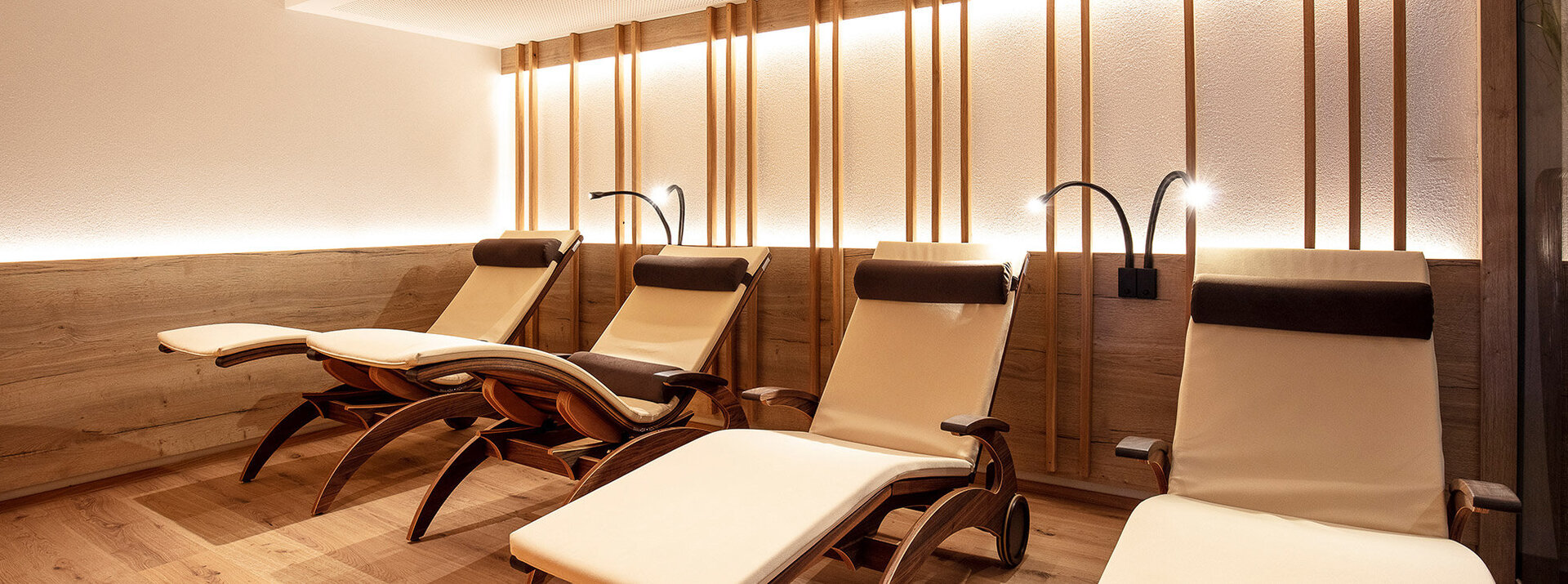 Ein entspannender Wellnessraum mit vier modernen, beigen Liegestühlen, Holzböden und -wänden sowie sanfter, warmer Beleuchtung. Jeder Liegestuhl verfügt über eine eigene Leseleuchte.