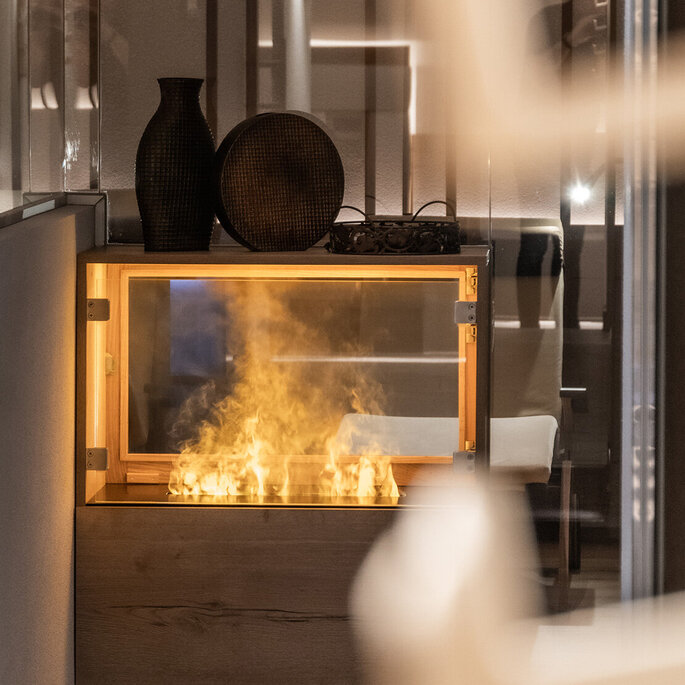 Ein kleiner, moderner Kamin mit sichtbaren Flammen ist in einen Holzschrank eingebaut. Darauf stehen zwei dunkle Ziervasen, und die Szene strahlt eine gemütliche, zeitgemäße Atmosphäre aus.