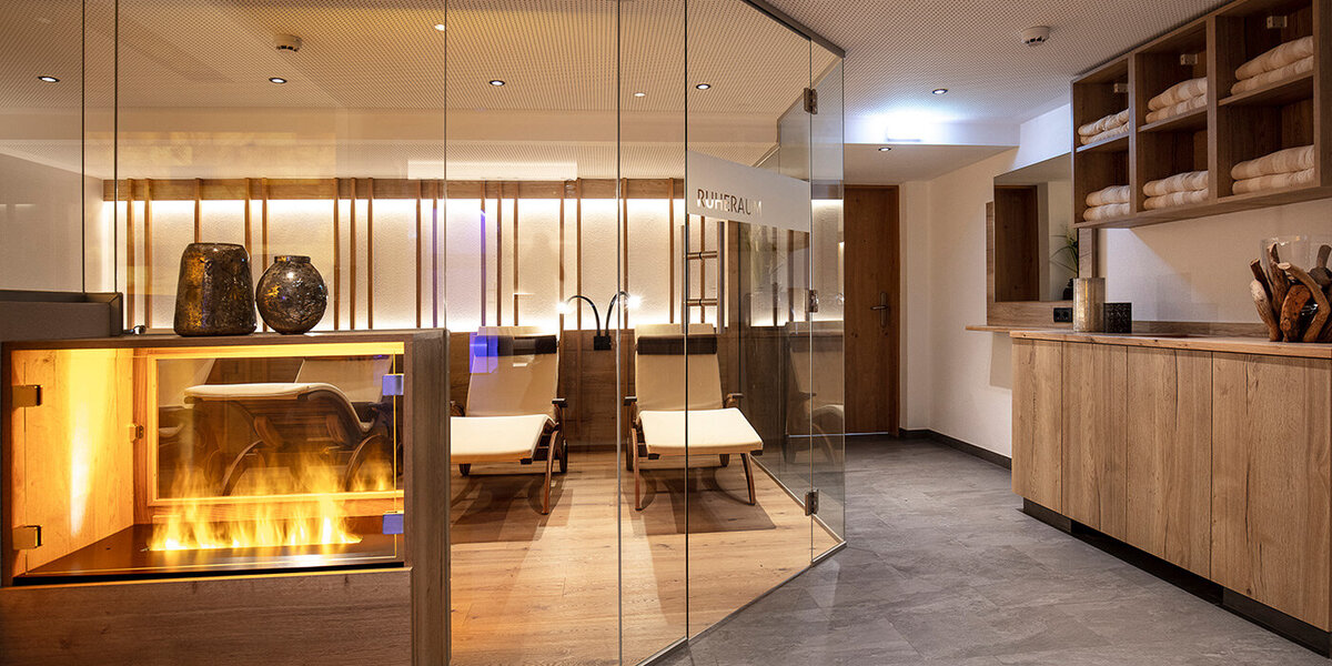 Moderner Wellnessbereich mit einer verglasten Sauna, Liegestühlen aus Holz, einem dekorativen Kamin und Regalen, auf denen gefaltete Handtücher gestapelt sind. Warme Beleuchtung sorgt für eine entspannte Atmosphäre.