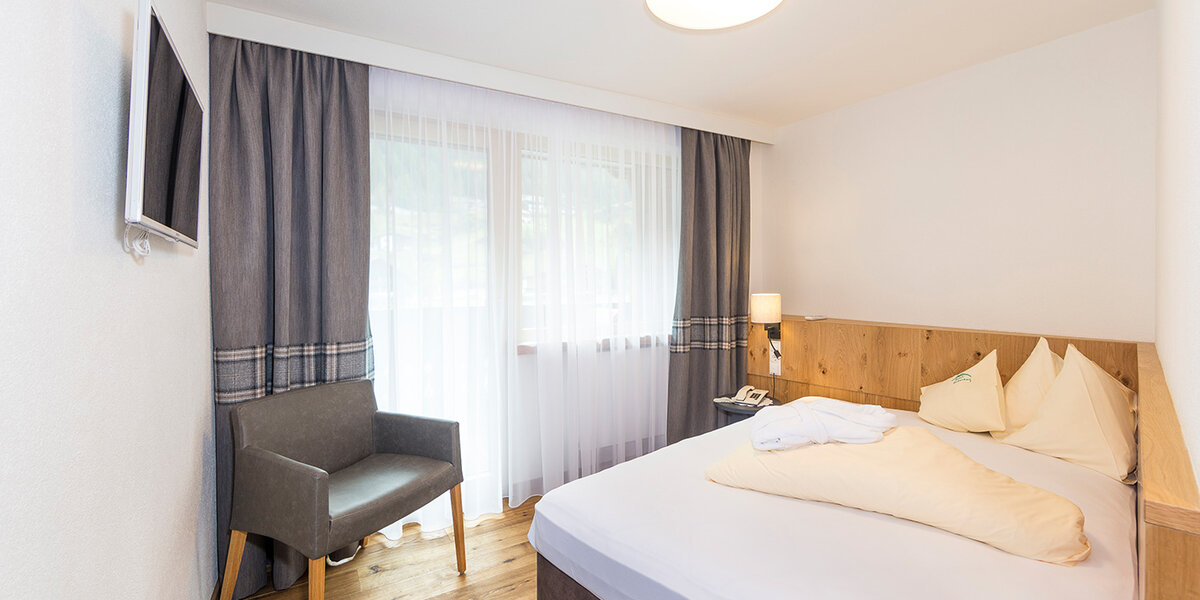 Modernes Hotelzimmer mit einem Doppelbett, einem grauen Sessel, einem an der Wand montierten Fernseher, einem Holzboden und einem großen Fenster, das mit transparenten Vorhängen und dunklen Gardinen verdeckt ist.