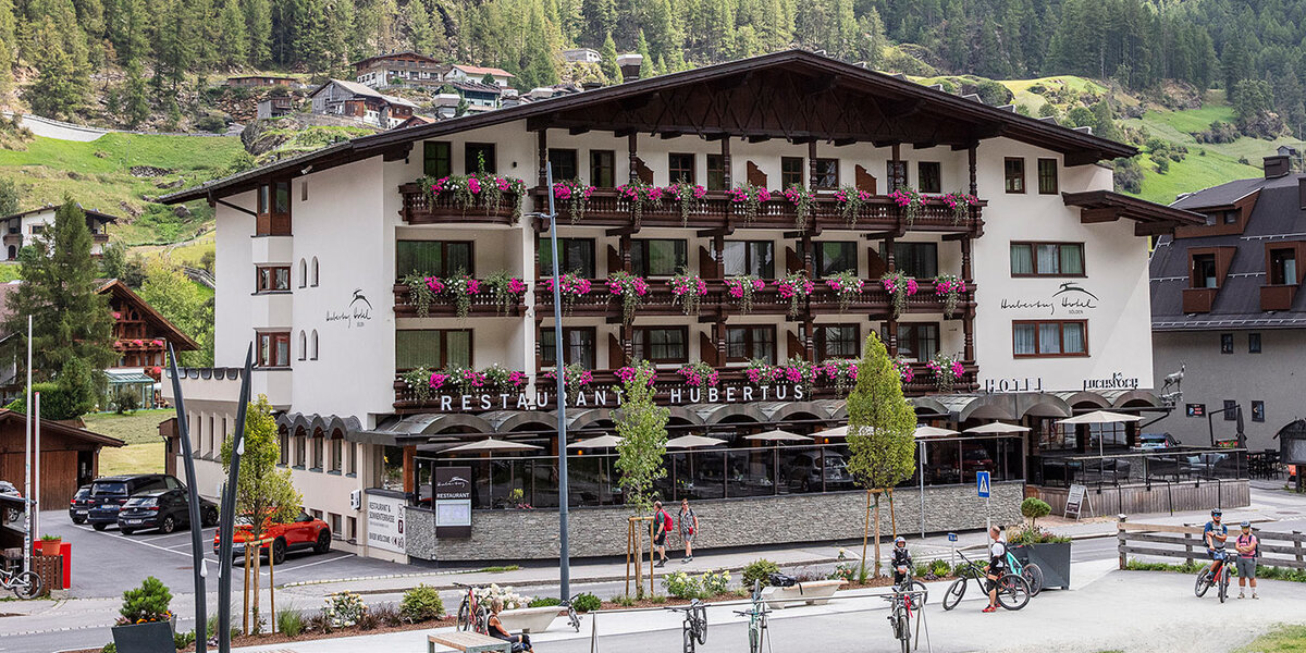 Ein vierstöckiges Hotel (Hubertus) mit Balkonen, die mit rosa Blumen geschmückt sind, in einem Bergdorf. Vor dem Haus stehen Fahrräder, und grüne, bewaldete Hügel umgeben die Gegend.