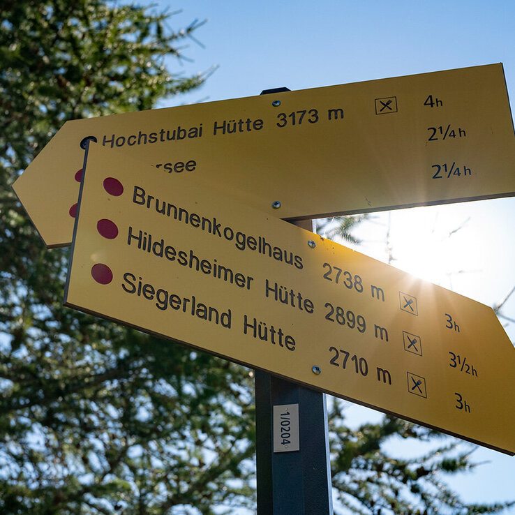 Gelbes Wanderzeichen auf Deutsch, das Wegbeschreibungen und Entfernungen zu Wanderhütten wie der Hochstubai-Hütte, dem Brunnenkogelhaus, der Hildesheimer Hütte und der Siegerland-Hütte anzeigt, im Hintergrund scheint die Sonne.