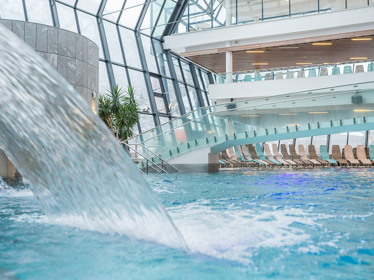 Ein Hallenbad mit einem kleinen Wasserfall, Liegestühlen, die am Rand aufgereiht sind, großen Glasfenstern und einer modernen Brücke, die über das Wasser führt.
