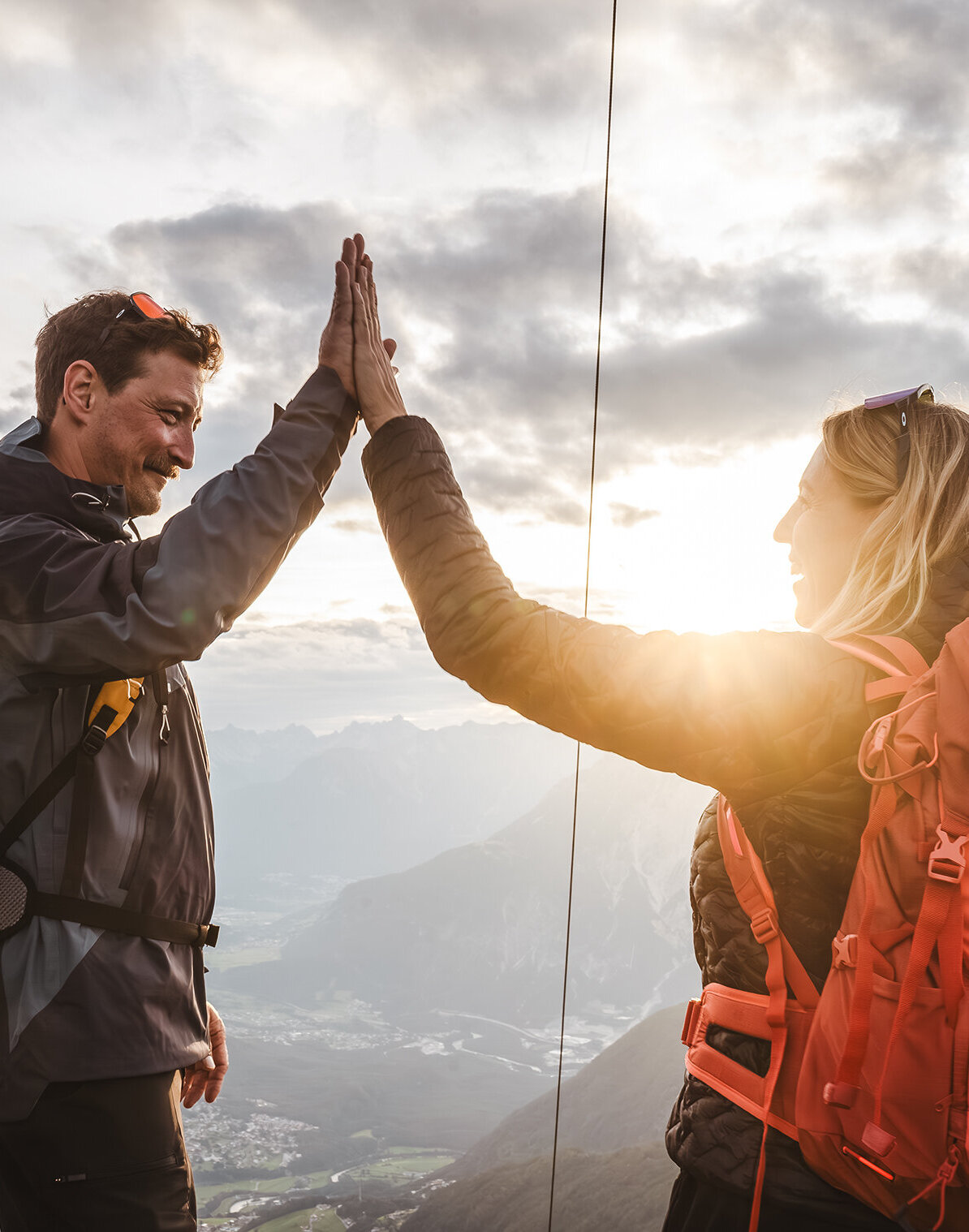 Zwei Wanderer mit Rucksäcken geben sich neben einem Holzpfahl auf einem Berggipfel ein High-Five, während im Hintergrund die Sonne scheint und malerische Wolken zu sehen sind.