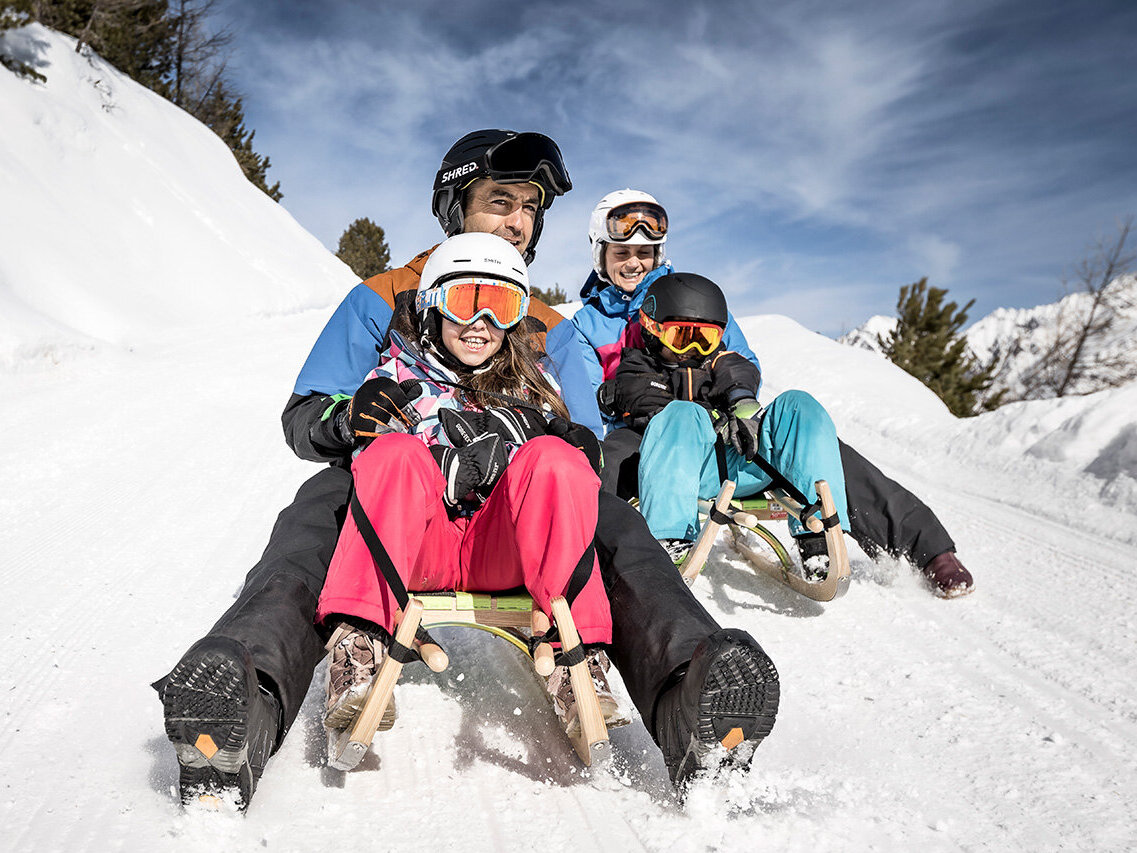 Drei Personen mit Helmen und Winterkleidung fahren mit der Rodel einen verschneiten Hügel hinunter, lächeln und genießen die Fahrt unter einem blauen Himmel mit einigen Wolken.