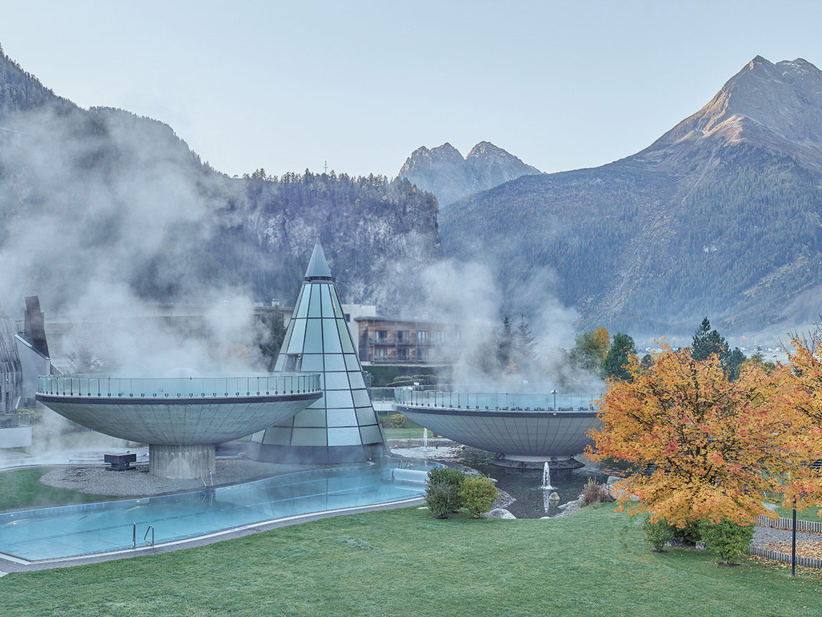 Ein modernes Spa mit zwei großen, dampfenden runden Becken und einer gläsernen Pyramidenkonstruktion, das im Freien inmitten von Bergen und herbstlich gefärbten Bäumen liegt, während in der kühlen Luft Nebel aufsteigt.