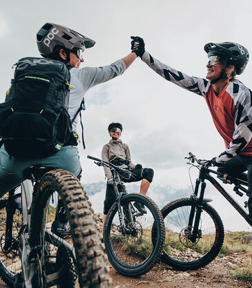 Drei Mountainbiker machen auf einem Trail im Freien eine Pause; zwei von ihnen geben sich lächelnd ein High Five, während der dritte Biker auf seinem Fahrrad sitzt und die Szene in entspannter Haltung beobachtet.