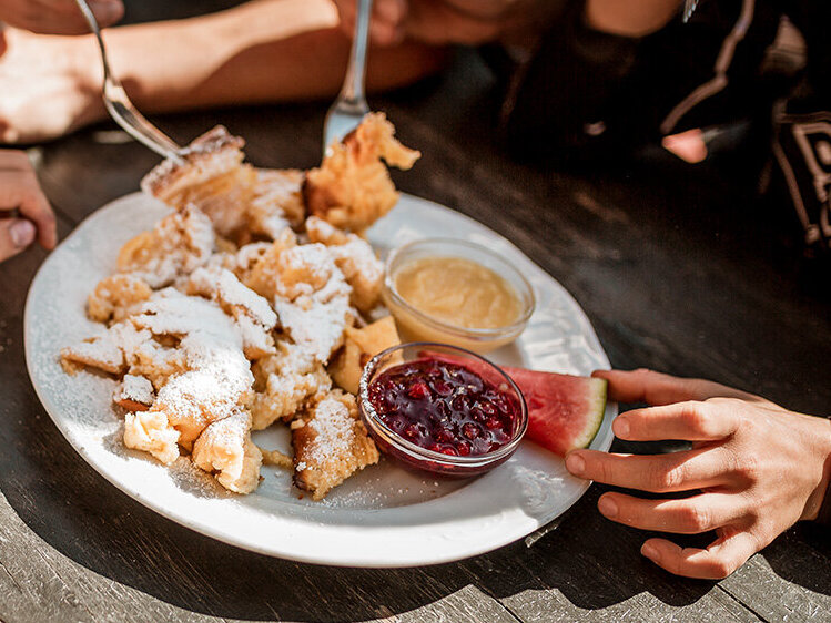 Vier Personen sitzen an einem Holztisch und teilen sich einen Teller mit Kaiserschmarren, der mit Puderzucker bestäubt ist, dazu stehen zwei Dip-Saucen – eine rote und eine gelbe – in kleinen Schälchen. Sonnenlicht taucht die Szene in helles Licht, und die Gruppe isst mit Gabeln.