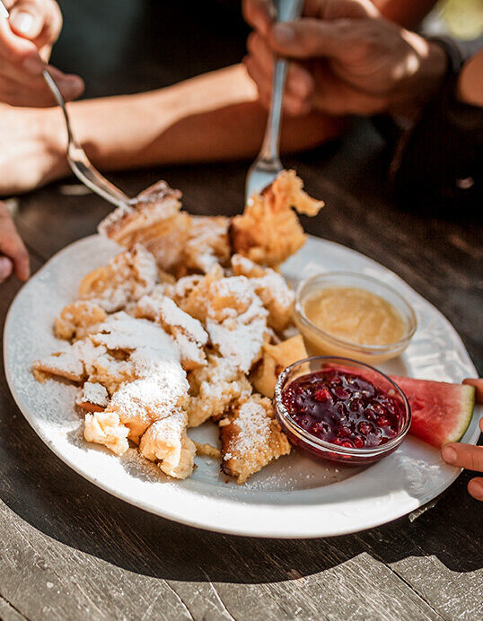 Vier Personen sitzen an einem Holztisch und teilen sich einen Teller mit Kaiserschmarren, der mit Puderzucker bestäubt ist, dazu stehen zwei Dip-Saucen – eine rote und eine gelbe – in kleinen Schälchen. Sonnenlicht taucht die Szene in helles Licht, und die Gruppe isst mit Gabeln.