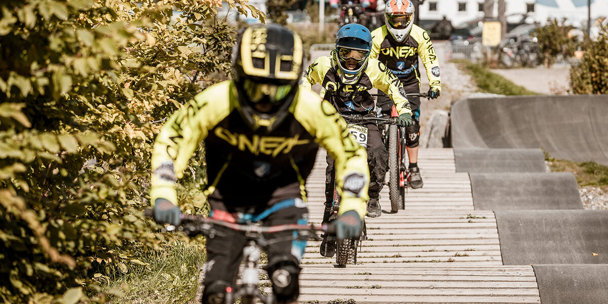 Drei Radfahrer in gelben Trikots und Helmen fahren mit Mountainbikes auf einem Holzsteg, umgeben von viel Grün; im unscharfen Hintergrund sind weitere Radfahrer und Gebäude zu sehen.