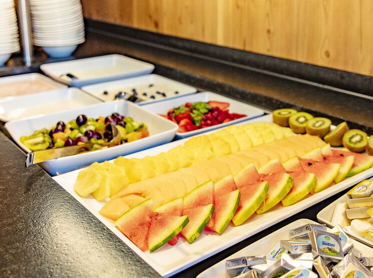 Auf einer Theke steht ein Buffet mit geschnittener Wassermelone, Cantaloupe-Melone, Ananas, Kiwi, gemischtem Obst und kleinen Butterpäckchen. Im Hintergrund sind Teller und Schüsseln gestapelt.