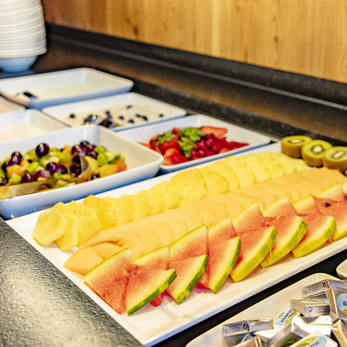 Auf einer Theke steht ein Buffet mit geschnittener Wassermelone, Cantaloupe-Melone, Ananas, Kiwi, gemischtem Obst und kleinen Butterpäckchen. Im Hintergrund sind Teller und Schüsseln gestapelt.