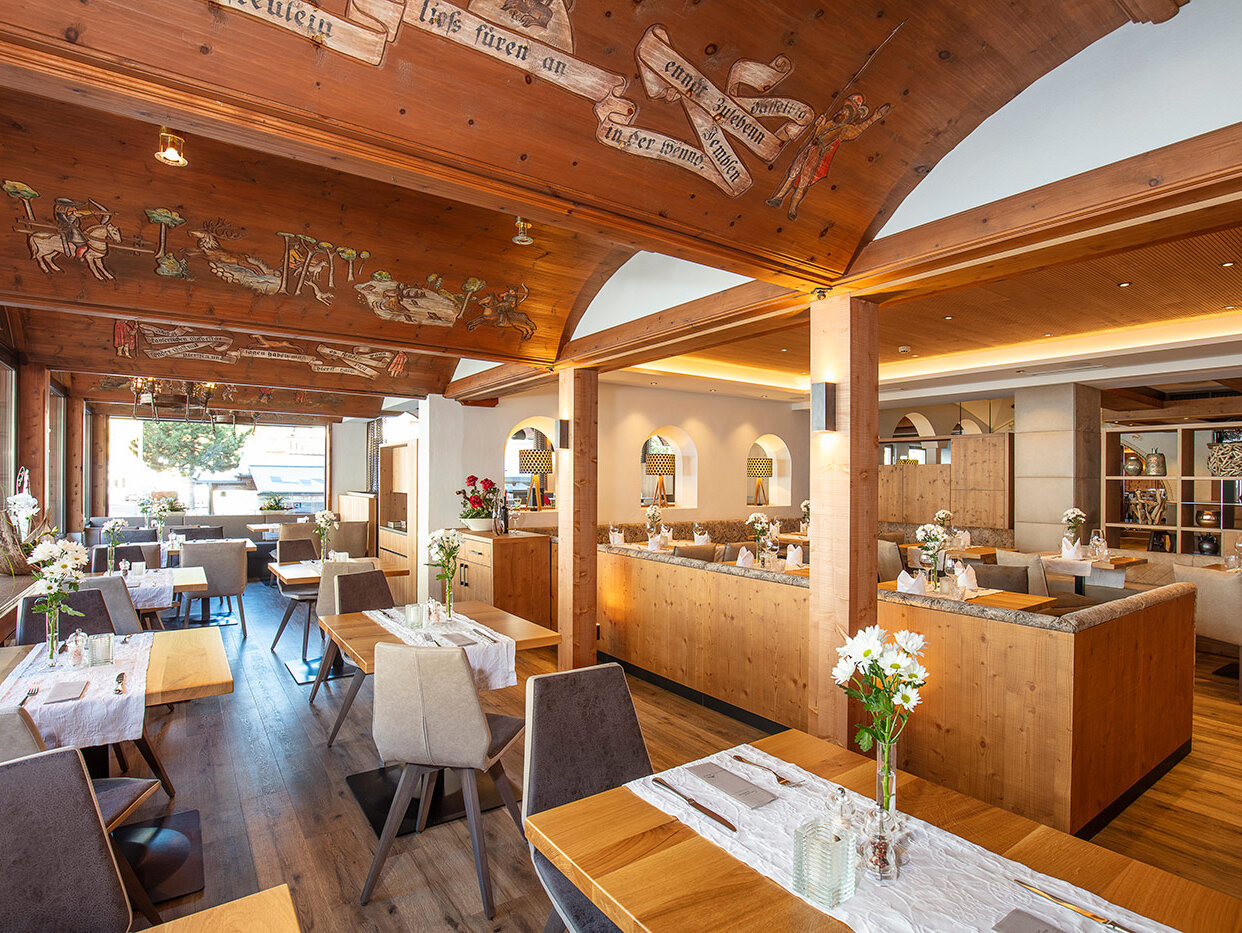 Ein helles, gemütliches Restaurantinterieur mit Holztischen, hellgrauen Stühlen, Blumen auf den Tischen und dekorativen Holzdecken mit gemalten Motiven. Große Fenster lassen Tageslicht herein.