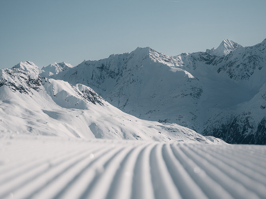 Eine frisch präparierte Skipiste mit parallelen Spuren im Schnee, die unter einem strahlend blauen Himmel auf die schneebedeckten Berge zuführt.