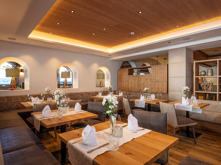 Ein gemütlicher Restaurantbereich mit Holztischen, braunen Stühlen, weißen Servietten und Blumengestecken als Tischdekoration. Warme Beleuchtung und dekorative Regale sorgen für eine einladende Atmosphäre.