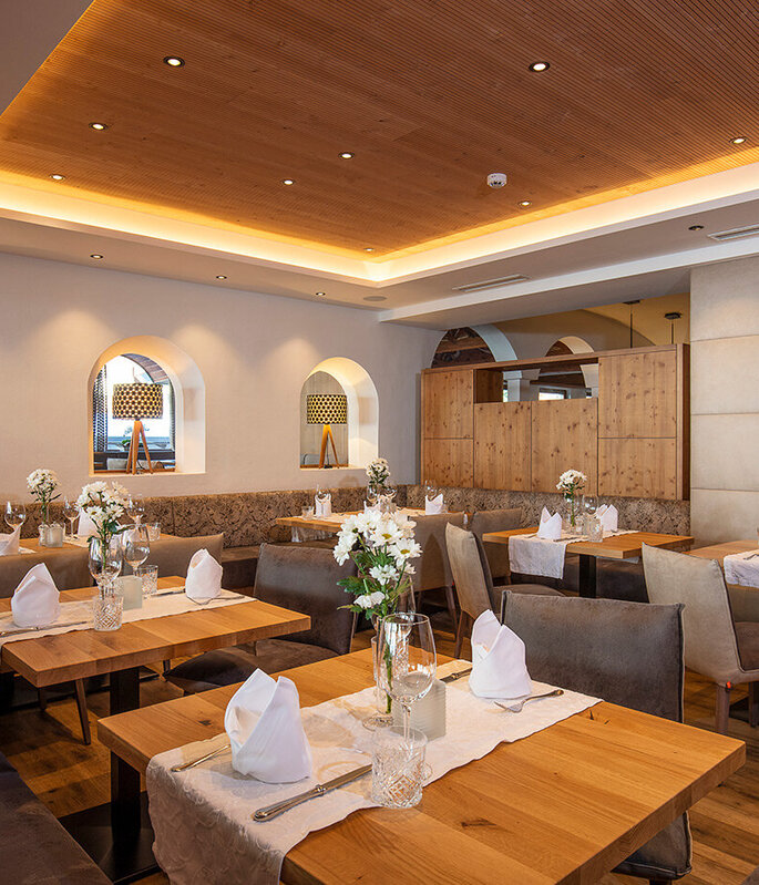 Ein gemütlicher Restaurantbereich mit Holztischen, braunen Stühlen, weißen Servietten und Blumengestecken als Tischdekoration. Warme Beleuchtung und dekorative Regale sorgen für eine einladende Atmosphäre.