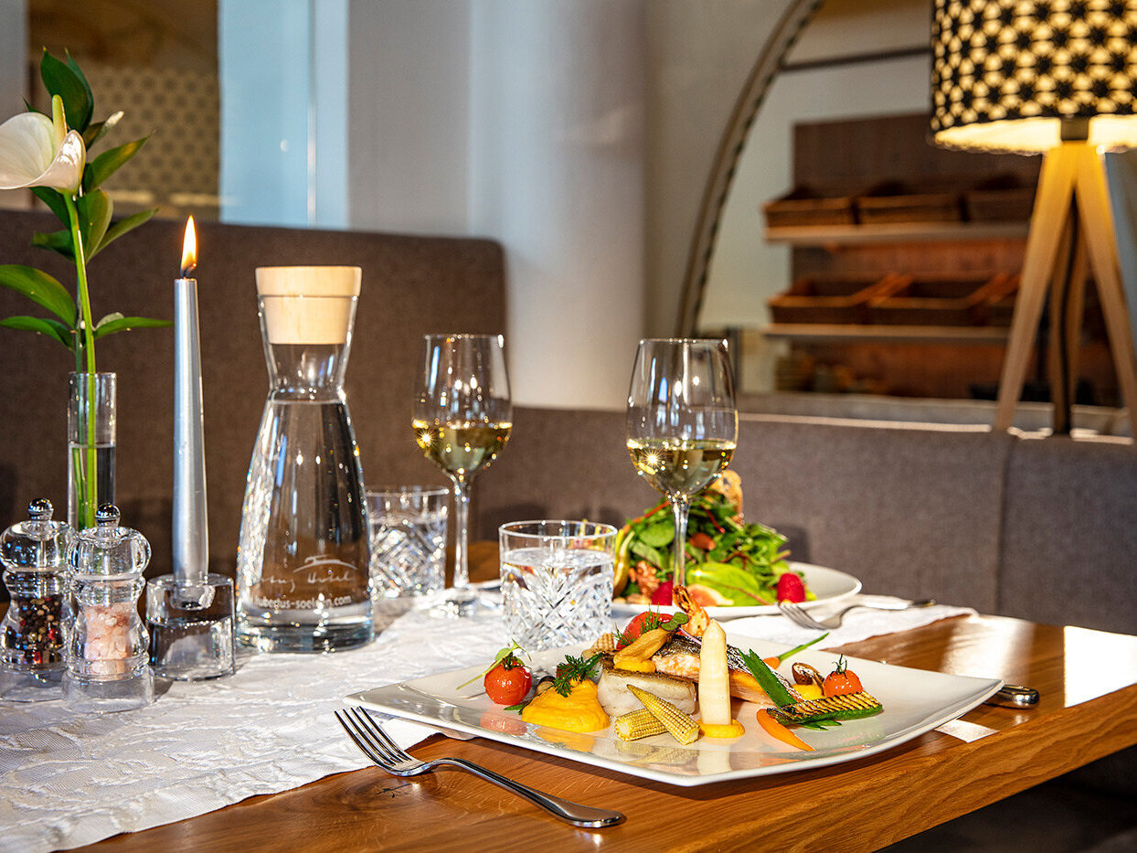 Ein für zwei Personen gedeckter Restauranttisch mit Weingläsern, einer Kerze, Wasser und Tellern mit Gourmetgerichten, darunter Fisch und Gemüse, auf einem Holztisch mit eleganter Dekoration.