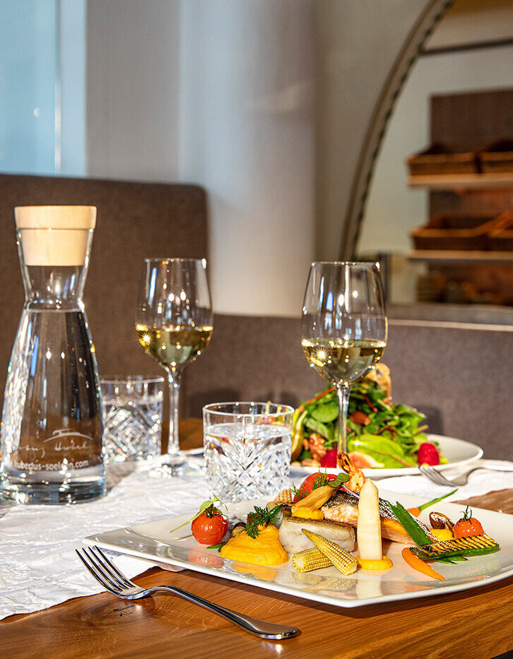 Ein für zwei Personen gedeckter Restauranttisch mit Weingläsern, einer Kerze, Wasser und Tellern mit Gourmetgerichten, darunter Fisch und Gemüse, auf einem Holztisch mit eleganter Dekoration.