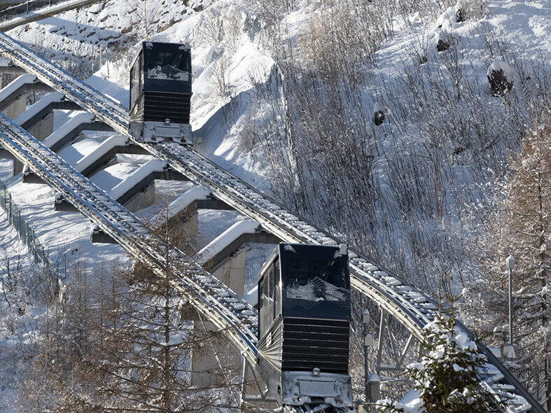 Unter strahlend blauem Himmel fährt eine steile Standseilbahn einen verschneiten, bewaldeten Berghang hinauf, wobei mehrere Wagen auf der Strecke hin und her fahren.