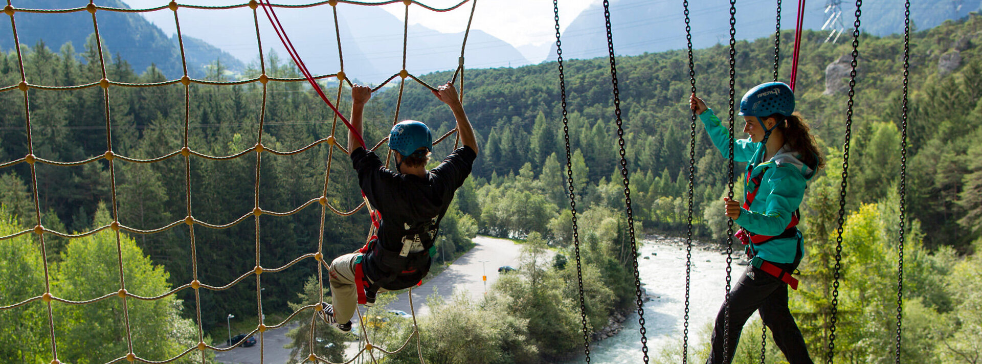 Zwei Personen, die Helme und Klettergurte tragen, bewegen sich auf einem Hochseilgarten über einem Fluss, umgeben von üppig grünen Bäumen und Bergen im Hintergrund.