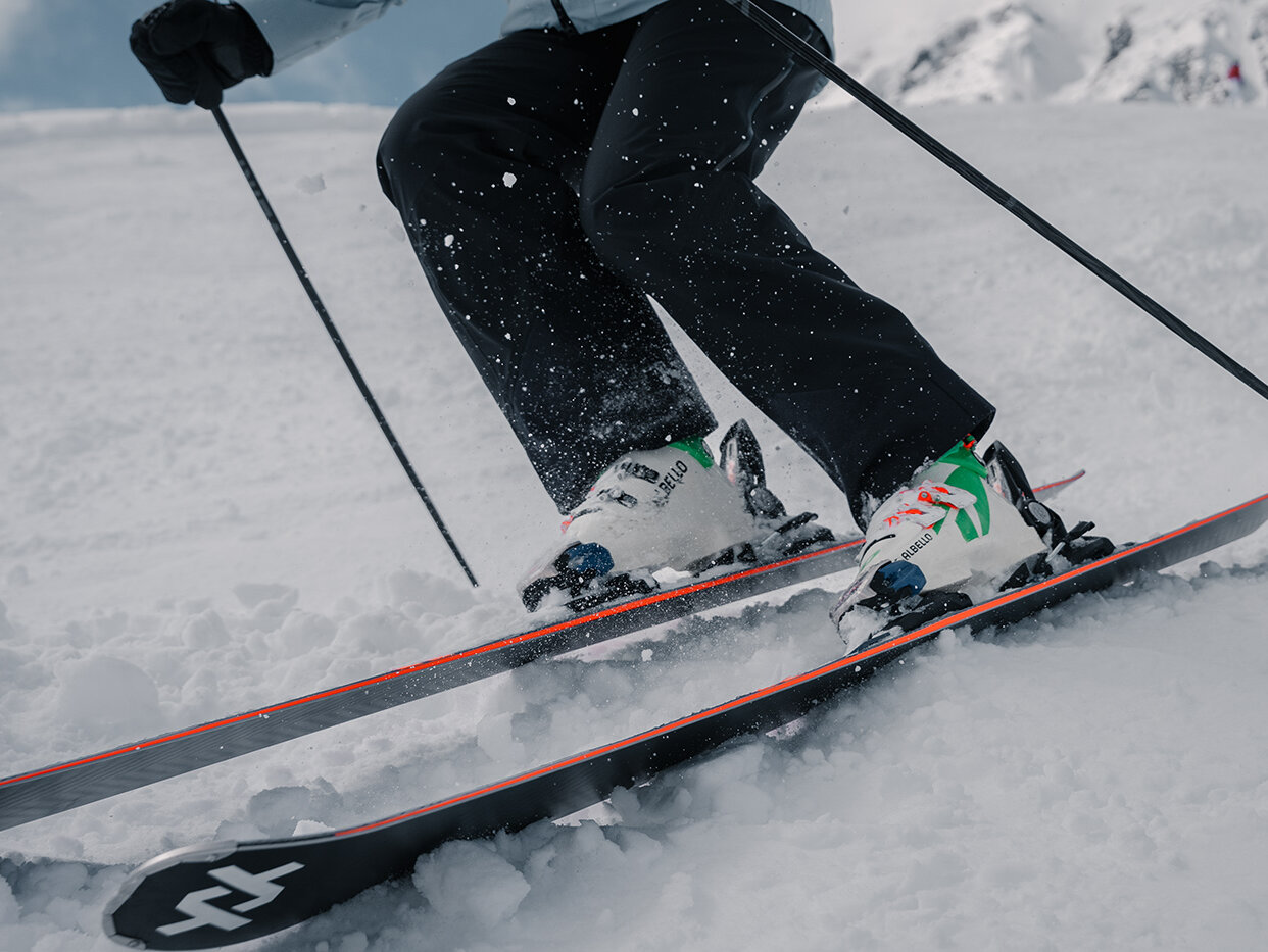 Nahaufnahme der Beine eines Skifahrers, der durch Neuschnee gleitet; er trägt eine schwarze Hose und weiße Skischuhe, die Skistöcke sind zu sehen, und um die Skier herum wirbelt Schnee auf.