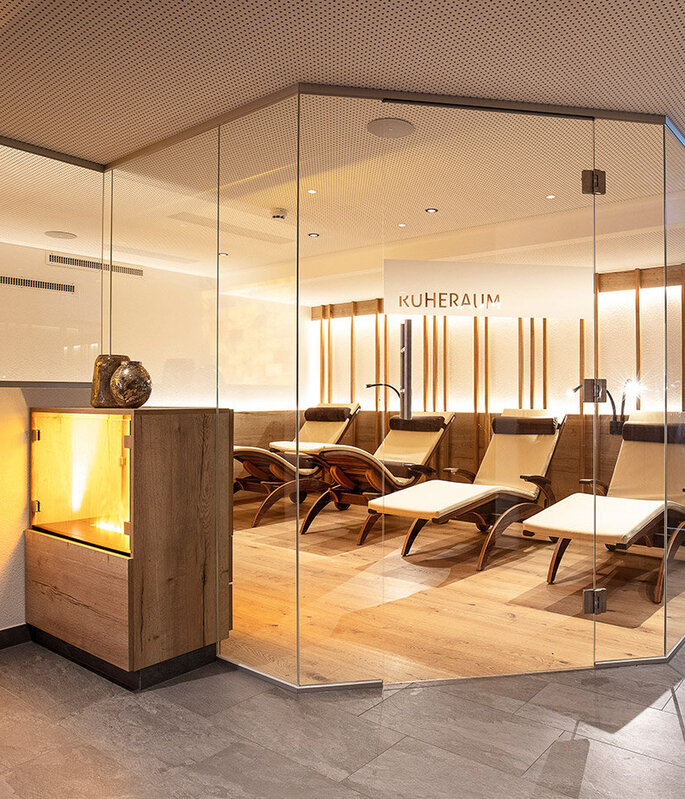 Eine moderne Entspannungslounge mit mehreren Liegesesseln hinter Glaswänden, warmer Beleuchtung, Holzböden und einem dekorativen Kamin, die eine gemütliche und einladende Atmosphäre schafft.