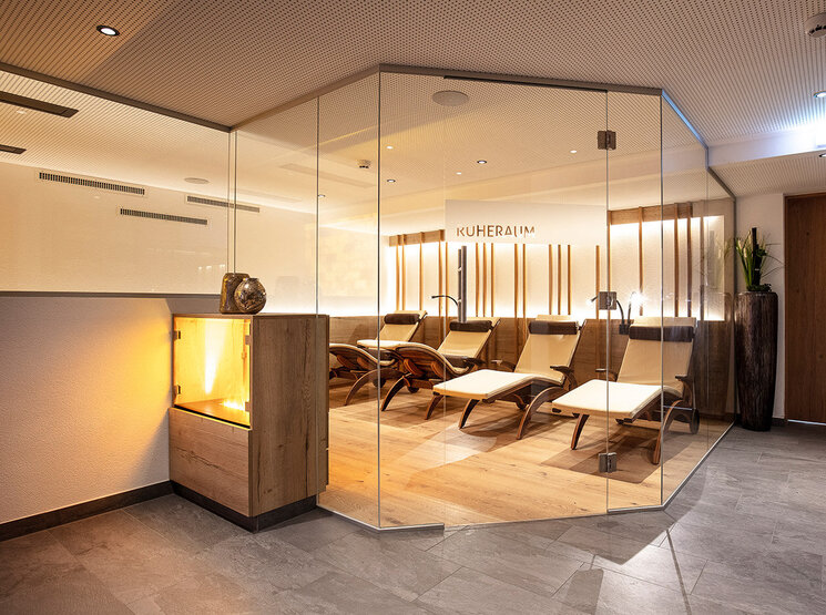 Eine moderne Entspannungslounge mit mehreren Liegesesseln hinter Glaswänden, warmer Beleuchtung, Holzböden und einem dekorativen Kamin, die eine gemütliche und einladende Atmosphäre schafft.