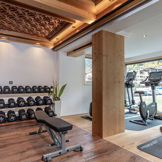 Ein moderner Fitnessraum mit einem Hantelständer, einer Trainingsbank und Cardiogeräten in der Nähe großer Fenster. Sonnenlicht strömt herein, und Holzakzente zieren die Decke und den Boden.
