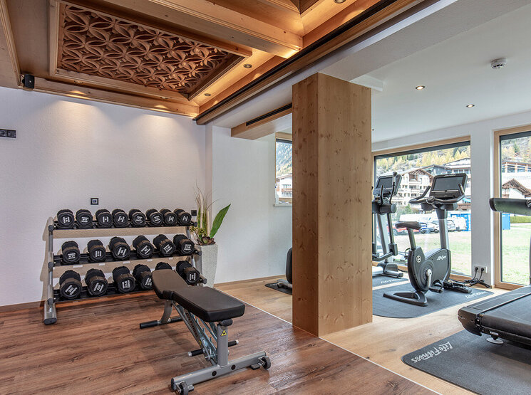 Ein moderner Fitnessraum mit einem Hantelständer, einer Trainingsbank und Cardiogeräten in der Nähe großer Fenster. Sonnenlicht strömt herein, und Holzakzente zieren die Decke und den Boden.