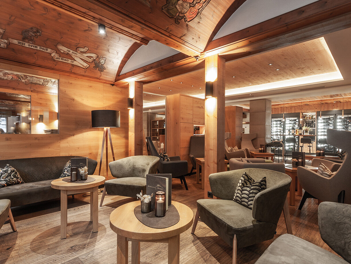 Gemütliche Lounge mit Holzwänden und -decke, modernen Sesseln, Sofas, kleinen runden Tischen und warmer Beleuchtung. Im Hintergrund sind eine Bar und ein Weinkeller zu sehen, die eine einladende Atmosphäre schaffen.
