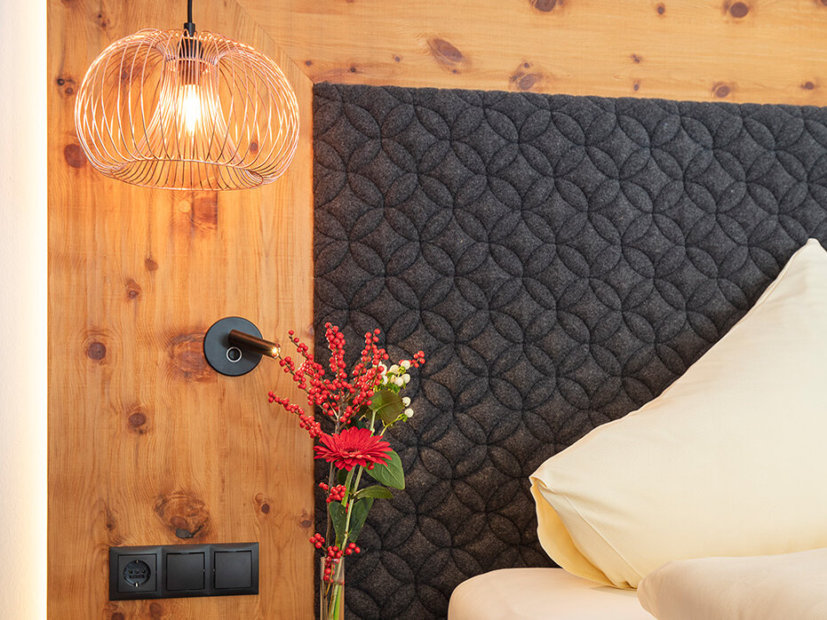 Eine moderne Hotelzimmer-Ecke mit einem Kopfteil aus Holz, einer gesteppten dunkelfarbigen Stoffverkleidung, einer Hängeleuchte, einem kleinen Nachttisch, einer Vase mit roten Blumen und ordentlich angeordneten cremefarbenen Kissen auf dem Bett.