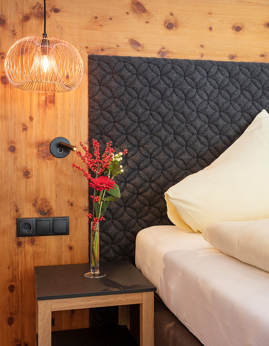 Eine moderne Hotelzimmer-Ecke mit einem Kopfteil aus Holz, einer gesteppten dunkelfarbigen Stoffverkleidung, einer Hängeleuchte, einem kleinen Nachttisch, einer Vase mit roten Blumen und ordentlich angeordneten cremefarbenen Kissen auf dem Bett.