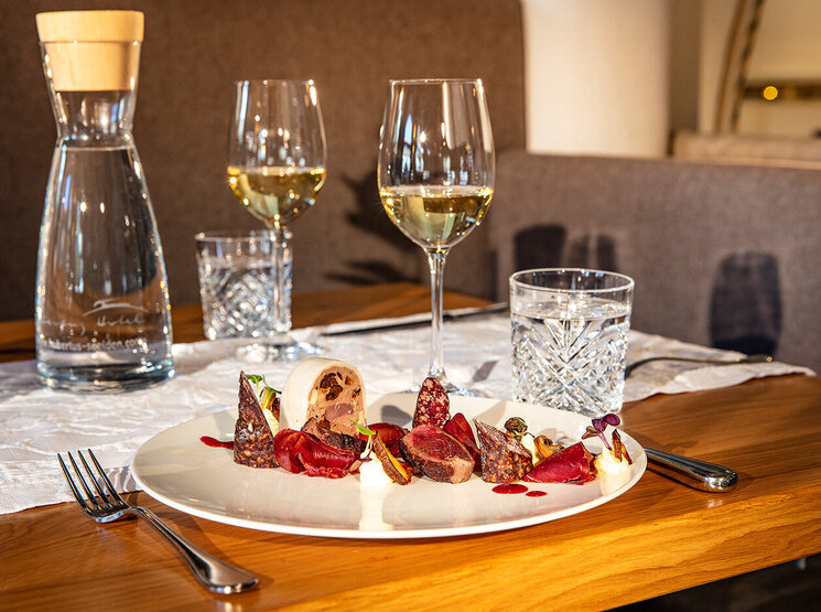 Auf einem Holztisch steht eine Platte mit verschiedenen Wurstwaren, Nüssen und Käse, dazu zwei Gläser Weißwein, Wassergläser, eine Karaffe und Besteck – alles für zwei Personen gedeckt.