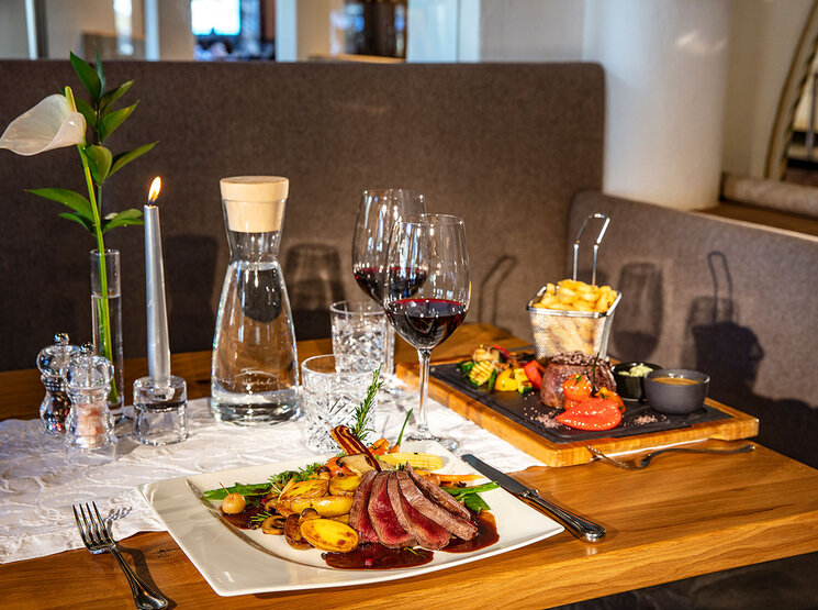 Ein Restauranttisch, gedeckt mit einem Gourmet-Menü bestehend aus Steak in Scheiben, Kartoffeln, Gemüse, Pommes frites, einer Kerze, einer Wasserkaraffe, Rotweingläsern und elegantem Geschirr auf einem Holztisch mit einer weißen Tischdecke.