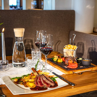 Ein Restauranttisch, gedeckt mit einem Gourmet-Menü bestehend aus Steak in Scheiben, Kartoffeln, Gemüse, Pommes frites, einer Kerze, einer Wasserkaraffe, Rotweingläsern und elegantem Geschirr auf einem Holztisch mit einer weißen Tischdecke.