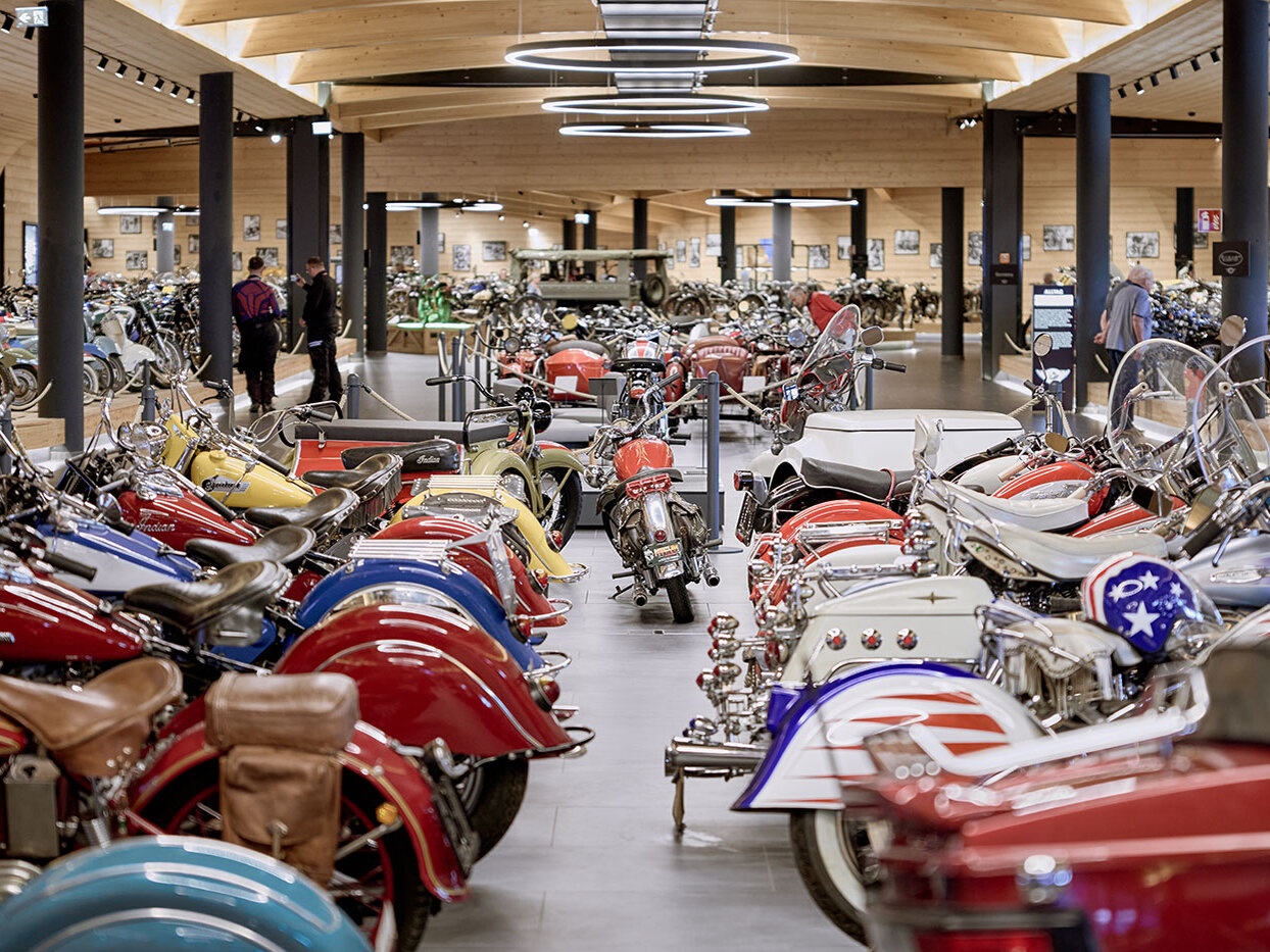 In einem geräumigen, modernen Museum sind Reihen klassischer und farbenfroher Motorräder ausgestellt, während einige Besucher umhergehen und sich die Exponate ansehen. An der Holzdecke hängen runde Lampen.