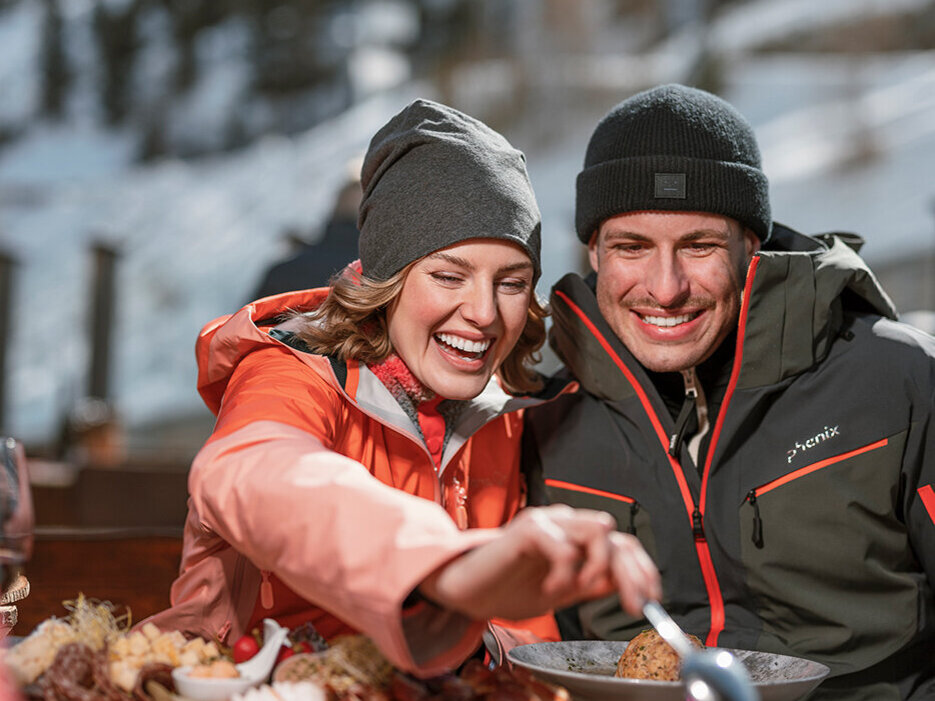 Zwei Menschen in Winterjacken und Mützen sitzen im Freien an einem verschneiten Ort in den Bergen, lächeln und essen gemeinsam an einem Tisch, auf dem Teller mit herzhaften Gerichten stehen.