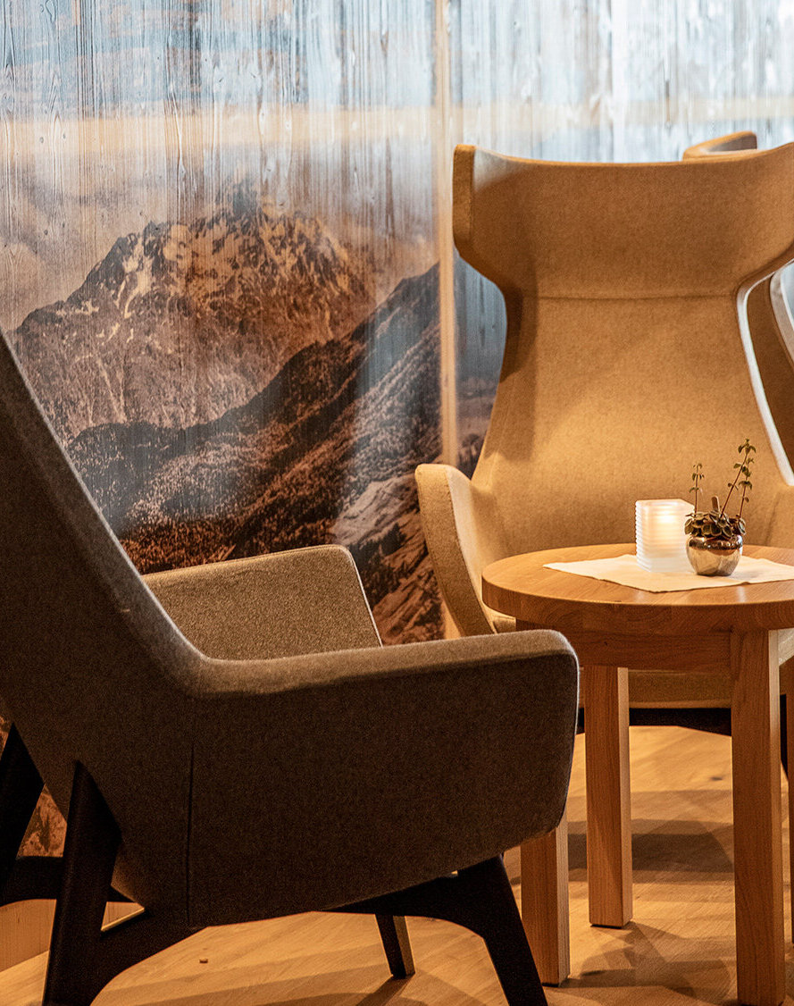 Eine gemütliche Lounge mit modernen Sesseln, die um einen kleinen Holztisch herum angeordnet sind, vor einer Wand mit einem malerischen Bergpanorama, sanft beleuchtet von Deckenstrahlern.