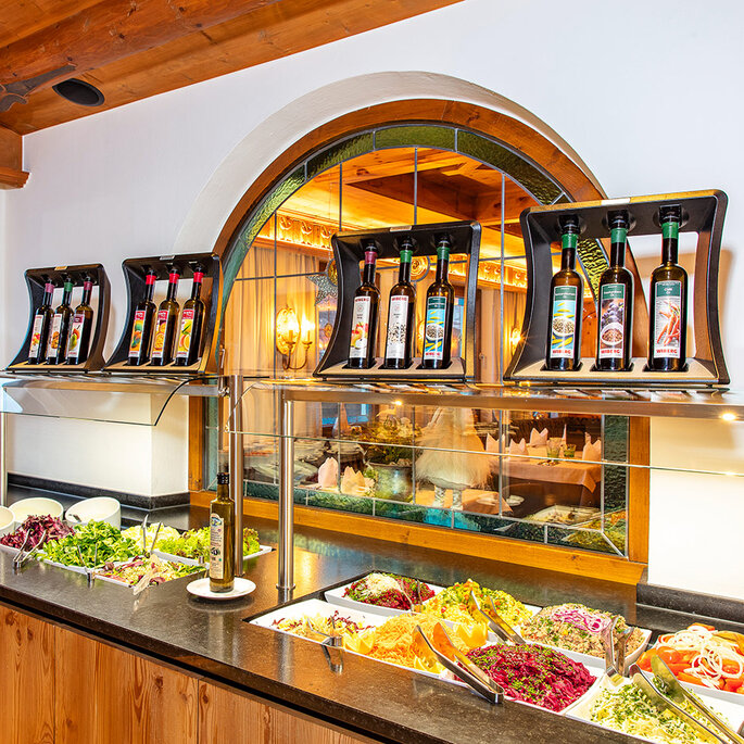 Eine Salatbar mit verschiedenen frischen Gemüsesorten und Blattgemüse, Tellern und Besteck auf der linken Seite sowie Flaschen mit Ölen oder Dressings, die in einem Regal über der Theke ausgestellt sind. Durch ein Fenster blickt man auf die Straße draußen.