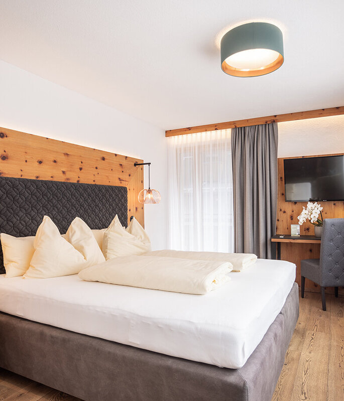 Ein modernes Hotelzimmer mit einem großen Bett, hellen Holzwänden, sanfter Beleuchtung, grauen Vorhängen, einem Schreibtisch mit Stuhl, einem an der Wand montierten Fernseher und einem großen Fenster mit weißen, durchsichtigen Vorhängen, durch die Tageslicht hereinströmt.