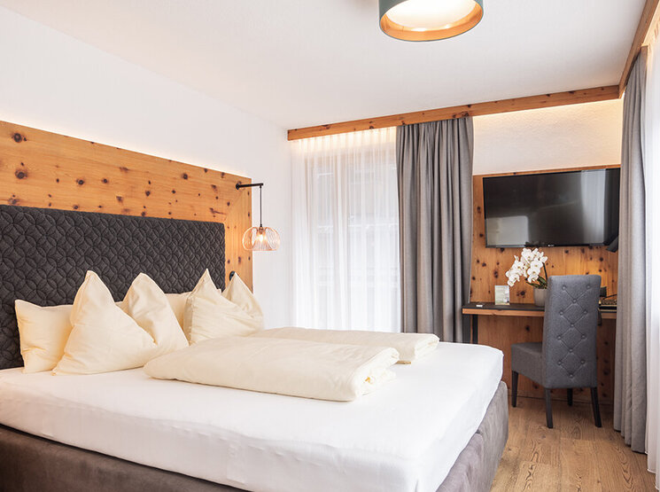 Ein modernes Hotelzimmer mit einem großen Bett, hellen Holzwänden, sanfter Beleuchtung, grauen Vorhängen, einem Schreibtisch mit Stuhl, einem an der Wand montierten Fernseher und einem großen Fenster mit weißen, durchsichtigen Vorhängen, durch die Tageslicht hereinströmt.