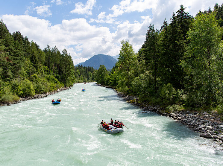 Gruppen von Menschen begeben sich auf eine Wildwasser-Rafting-Tour auf einem breiten, reißenden Fluss, der von dichtem, grünem Wald umgeben ist, während im Hintergrund unter einem teilweise bewölkten Himmel Berge zu sehen sind.