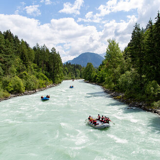 Gruppen von Menschen begeben sich auf eine Wildwasser-Rafting-Tour auf einem breiten, reißenden Fluss, der von dichtem, grünem Wald umgeben ist, während im Hintergrund unter einem teilweise bewölkten Himmel Berge zu sehen sind.